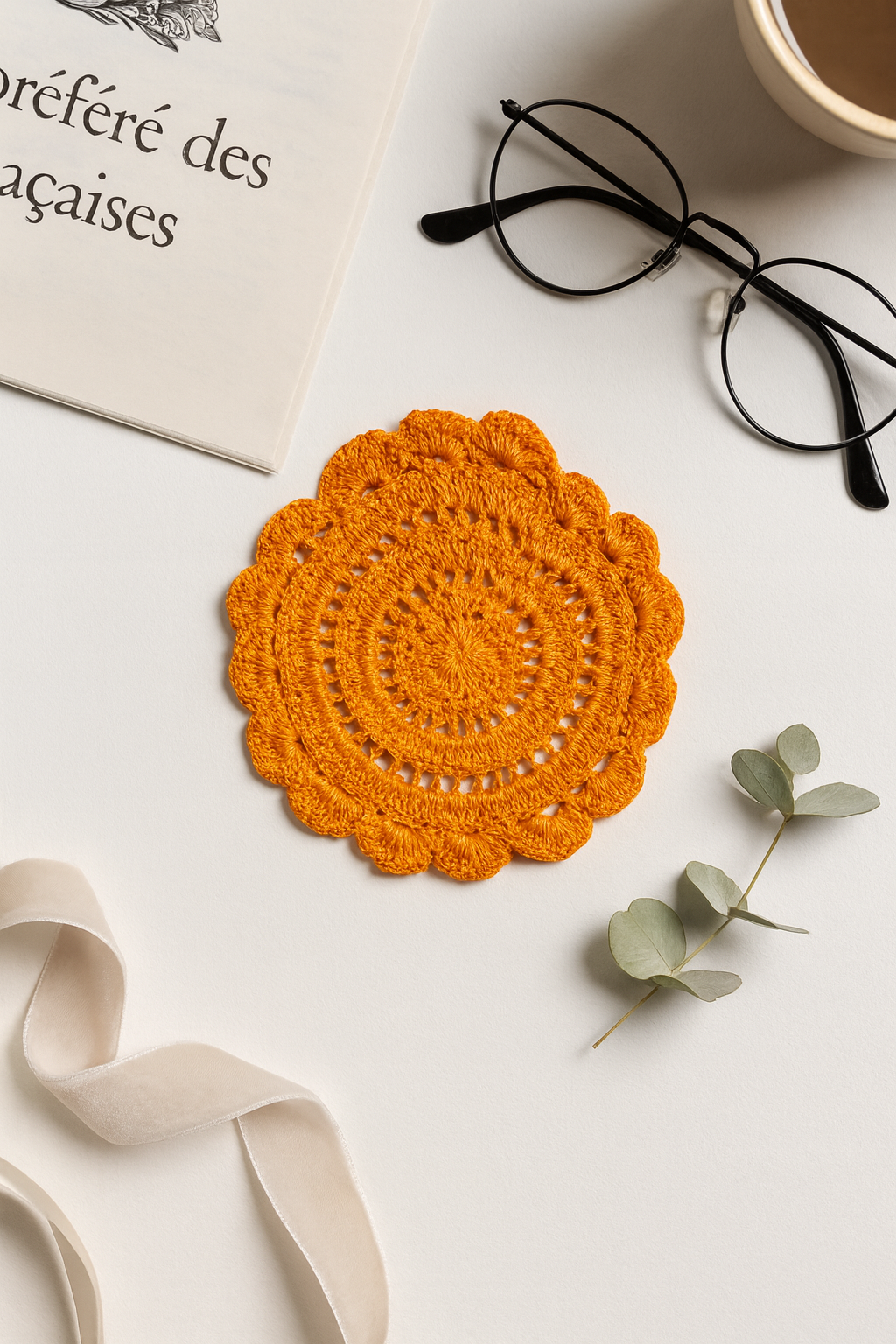 Orange crocheted coaster on a white surface with glasses, book, and eucalyptus leaves.