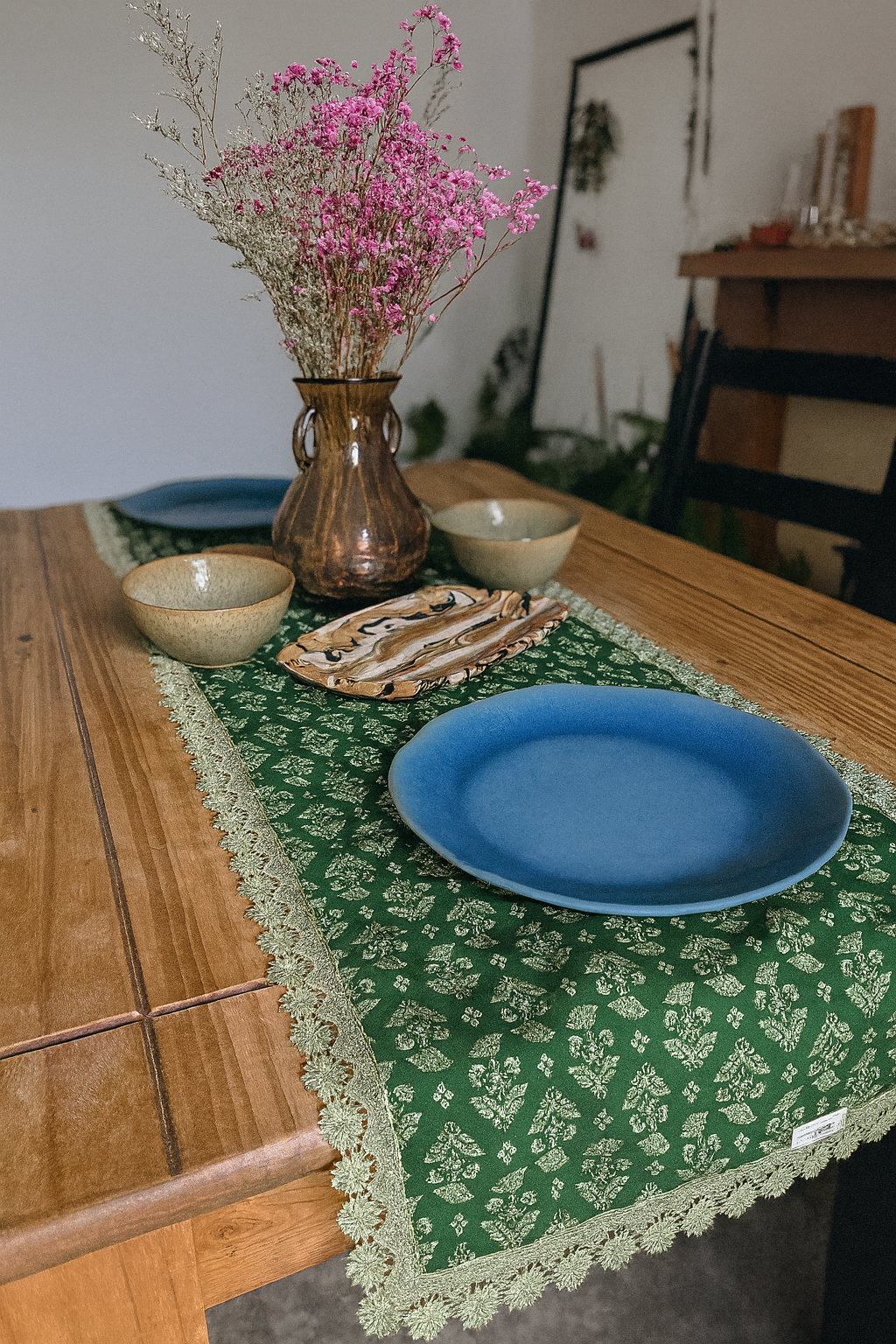Table setting with blue plates, a vase of pink flowers, and a green lace floral hand block printed tablecloth.