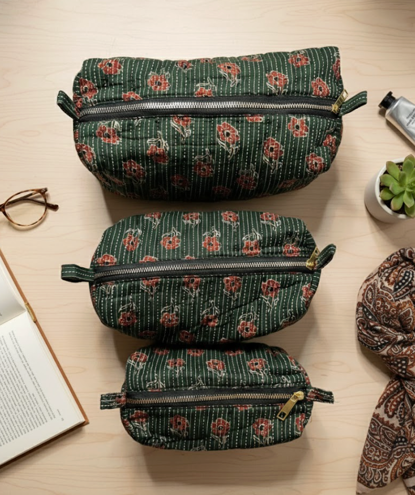 1 large, 1 medium and 1 small washer bag on a wooden table. Deside the bags are a book and spectacles on the left and and scarf, a small house plant and a handcream on right. 