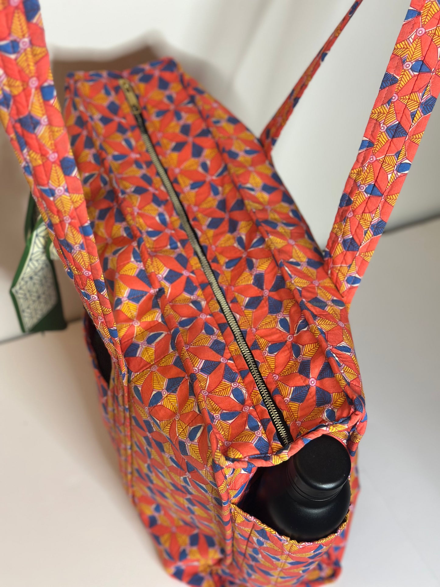a colourful patterned bag with it's zipper being highlighted, a black bottle in the side pocket on a white background