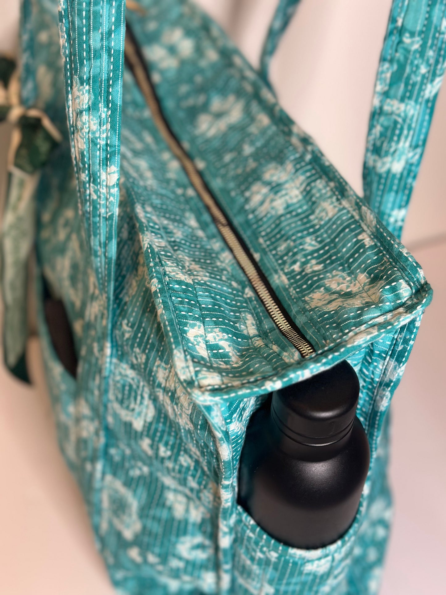 Teal patterned bag with a black bottle inside on a light pink background